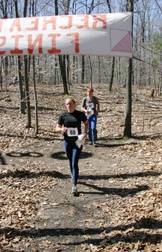 one orienteer runs under the finish banner in front of another